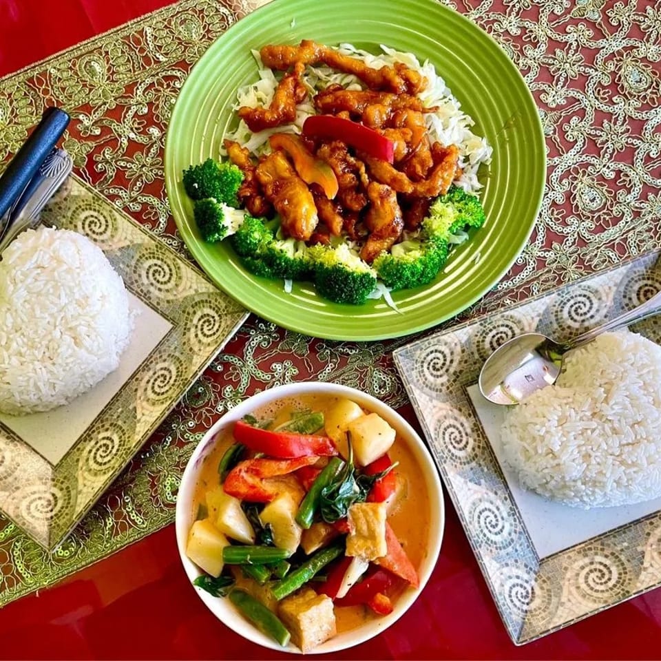 Three Thai dishes: crispy chicken, vegetable curry, and two heart-shaped servings of rice on a red tablecloth.